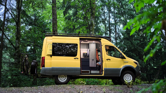 A yellow Nomad Vanz custom Sprinter conversion parked on a gravel forest trail with its sliding door open, revealing a clean grey interior with modular cabinetry — a mountain bike mounted on the rear hitch rack visible in the background.