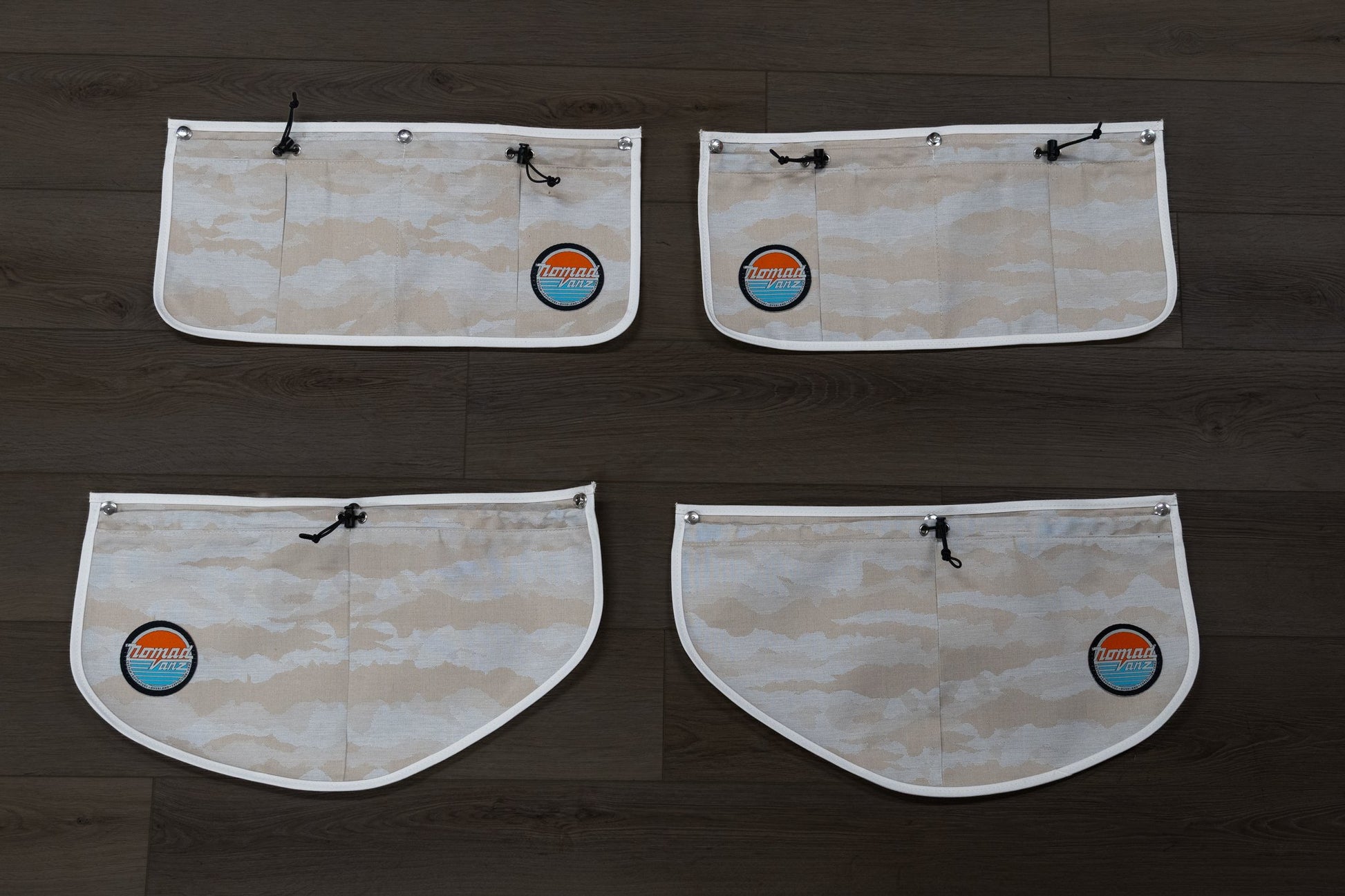 Back view of all four Nomad Vanz Rear Door Storage Pocket System pockets in Cirrus Sand camo laid flat on dark wood surface showing snap fastener attachment points, zippered openings, and Nomad Vanz logo patches