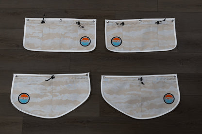 Back view of all four Nomad Vanz Rear Door Storage Pocket System pockets in Cirrus Sand camo laid flat on dark wood surface showing snap fastener attachment points, zippered openings, and Nomad Vanz logo patches