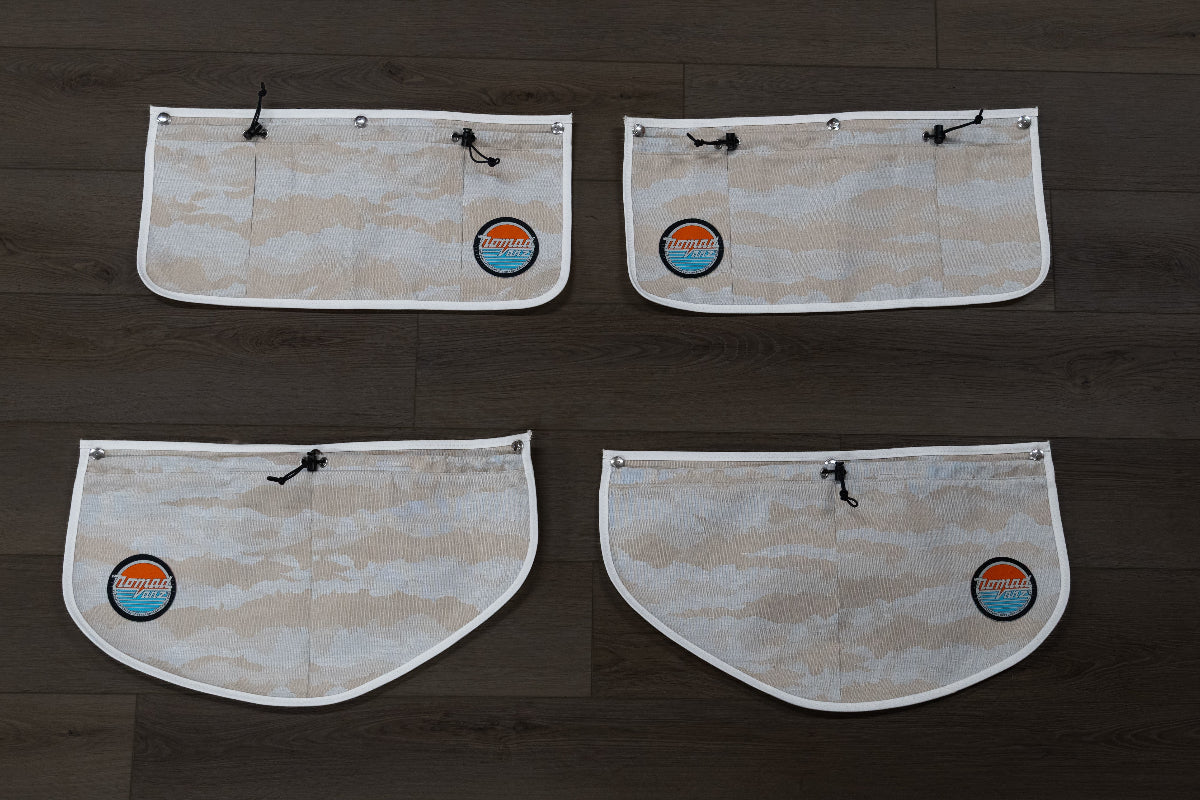 Back view of all four Nomad Vanz Rear Door Storage Pocket System pockets in Cirrus Sand camo laid flat on dark wood surface showing snap fastener attachment points, zippered openings, and Nomad Vanz logo patches