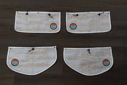 Back view of all four Nomad Vanz Rear Door Storage Pocket System pockets in Cirrus Sand camo laid flat on dark wood surface showing snap fastener attachment points, zippered openings, and Nomad Vanz logo patches