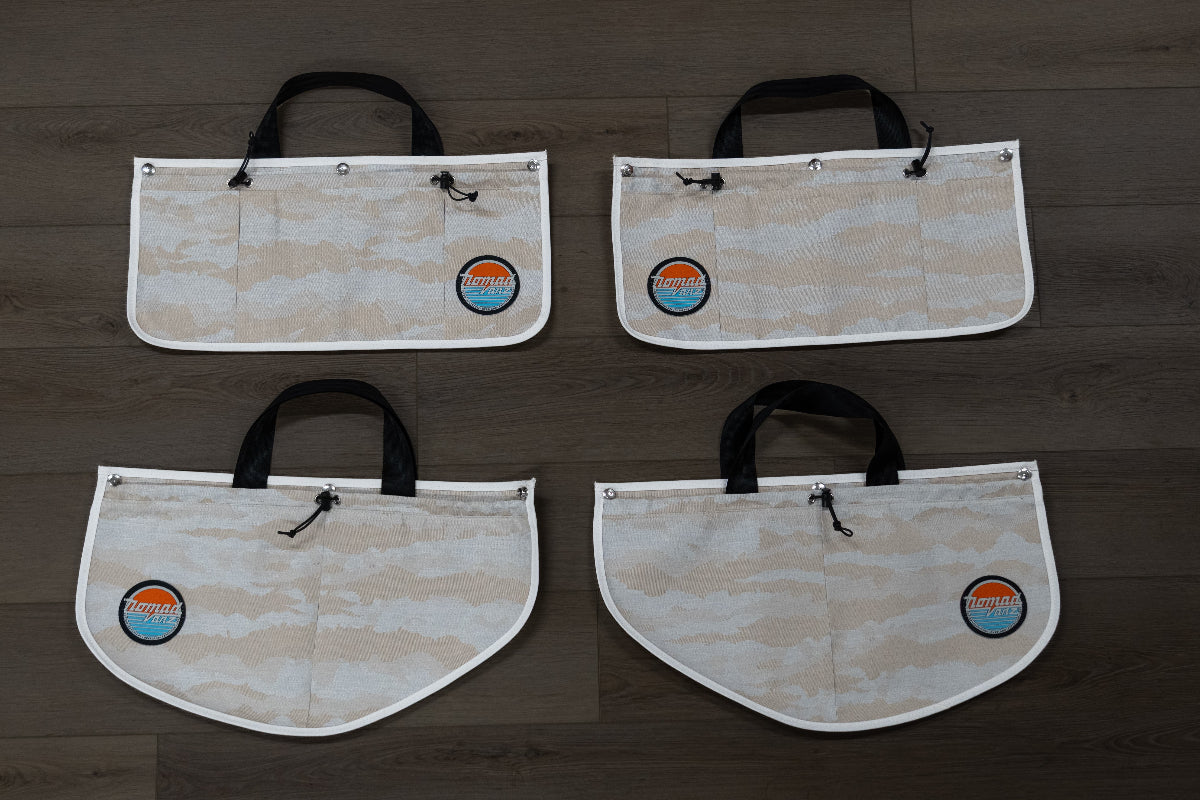 All four Nomad Vanz Rear Door Storage Pocket System pockets in Cirrus Sand camo laid flat on dark wood surface showing two rectangular upper pockets and two rounded lower pockets with carry handles, toggle closures, and Nomad Vanz logo patches