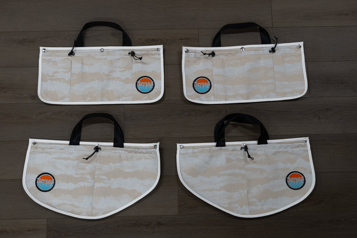 Front view of all four Nomad Vanz Rear Door Storage Pocket System pockets in Cirrus Sand camo laid flat on dark wood surface showing the carry handles, bungee drawstring closures, white piping trim, and Nomad Vanz logo patches