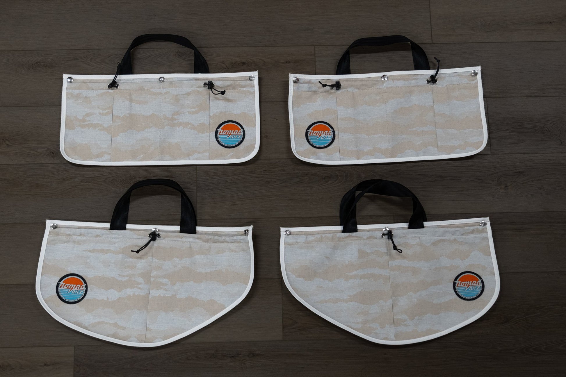Front view of all four Nomad Vanz Rear Door Storage Pocket System pockets in Cirrus Sand camo laid flat on dark wood surface showing the carry handles, bungee drawstring closures, white piping trim, and Nomad Vanz logo patches