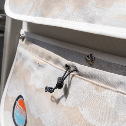 Macro detail of the Nomad Vanz Rear Door Storage Pocket System in Cirrus Sand camo showing the bungee drawstring cord closure, snap fastener, white piping trim, and Nomad Vanz logo patch on durable canvas fabric