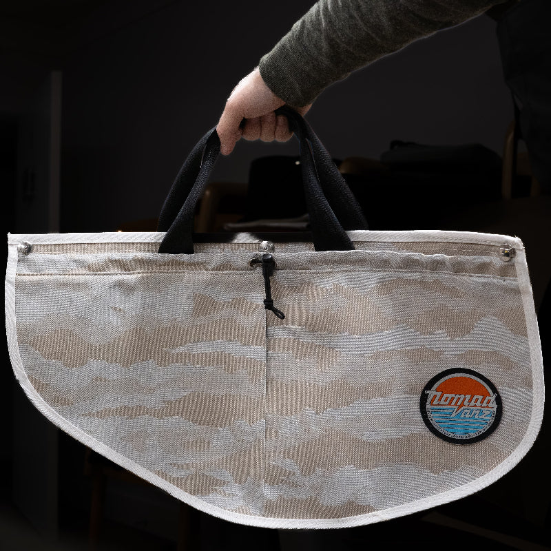 Person holding a Nomad Vanz Rear Door Storage Pocket in Cirrus Sand camo by its built-in carry handles, showing the removable design with toggle closure, snap fasteners, and Nomad Vanz logo patch