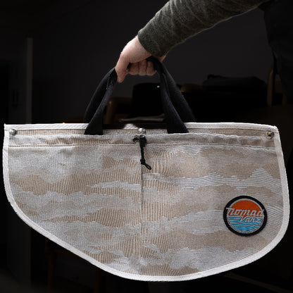 Person holding a Nomad Vanz Rear Door Storage Pocket in Cirrus Sand camo by its built-in carry handles, showing the removable design with toggle closure, snap fasteners, and Nomad Vanz logo patch