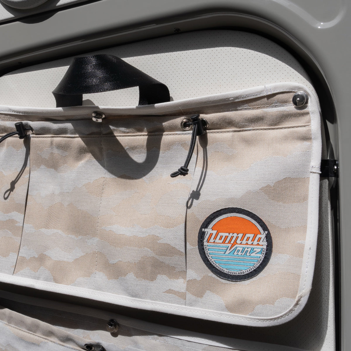 Close-up of Nomad Vanz Rear Door Storage Pocket in Cirrus Sand camo mounted on Sprinter rear door showing toggle closure, snap fastener hardware, carry handle, and Nomad Vanz logo patch