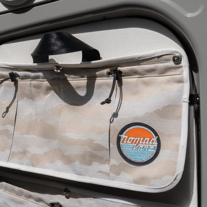 Close-up of Nomad Vanz Rear Door Storage Pocket in Cirrus Sand camo mounted on Sprinter rear door showing toggle closure, snap fastener hardware, carry handle, and Nomad Vanz logo patch