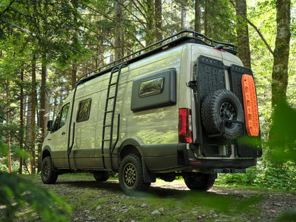 Side rear view of a Nomad Vanz Sprinter camper van in the forest showing full roof rack, rear ladder, spare tire, and traction board