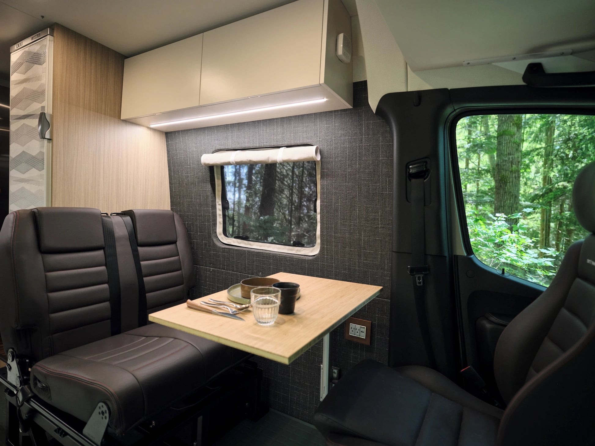 Nomad Vanz Sprinter interior dinette with bamboo table, leather bench seat, overhead cabinets, and a side window framing the forest