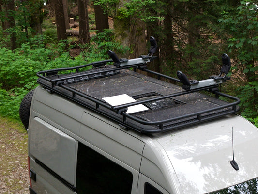 Elevated view of the roof rack with mesh deck and Thule kayak mounts on an olive Nomad Vanz Twoonie Sprinter camper van, with a spare tire and forest behind